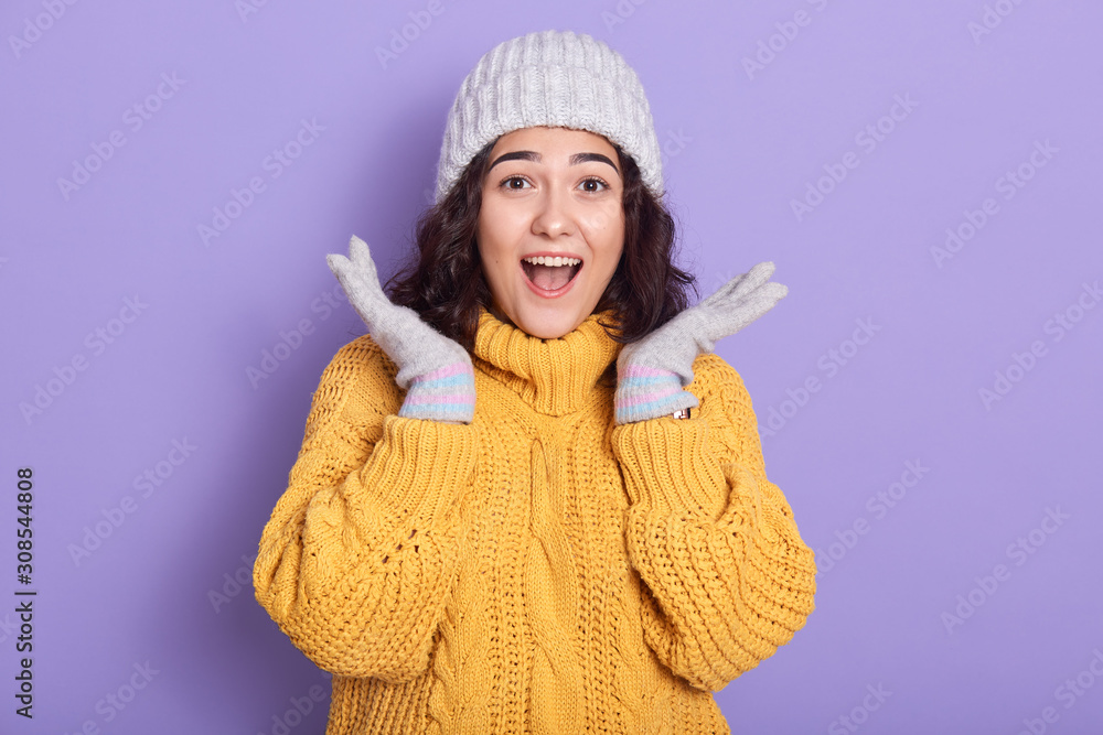 Image of shocked impressed beautiful young woman raising hands, opening mouth and eyes widely, wearing yellow sweater and white hat, gloves, standing isolated over lilac background in studio.