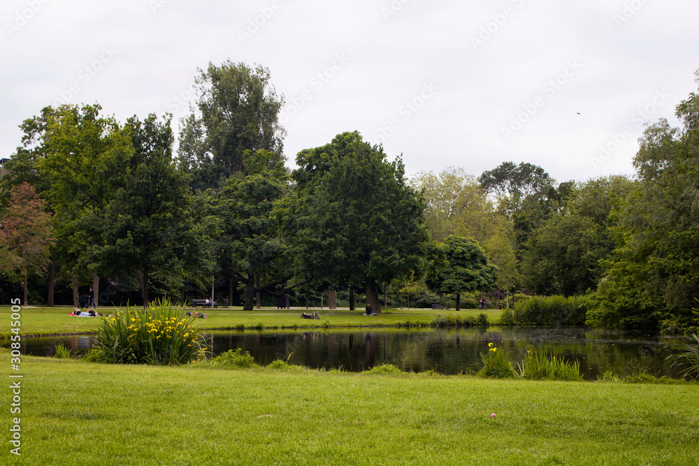 View of people hanging out, trees, grass fields and lake at Vondelpark in Amsterdam. It is a public urban park of 47 hectares. It is a summer day.