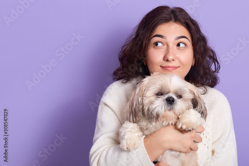 Image of beautiful attractive young female with black curly hair, looking aside, holding her small domestic animal Maltese dog in hands, spending time with pleasure. Copyspace for advertisement.