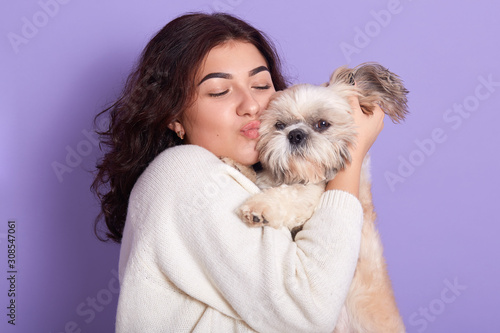 Horizontal picture of magnetic charming black haired woman with pet in her hands, hugging her domestic animal, kissing it with closed eyes, wearing white sweater, posing over lilac background.
