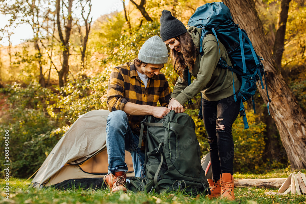 Young beautiful couple gather their backpacks on a hike. Attractive woman and handsome man relaxing together in nature near tent. Young beautiful couple gather their backpacks on a hike. Attractive woman and handsome man relaxing together in nature near tent.