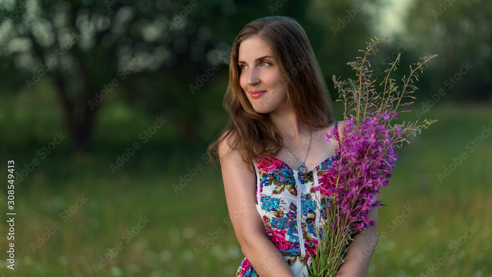 Beautiful girl in multicolored dress stands with purple flowers at sunset