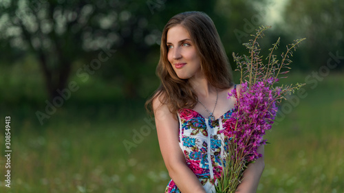 Beautiful girl in multicolored dress stands with purple flowers at sunset