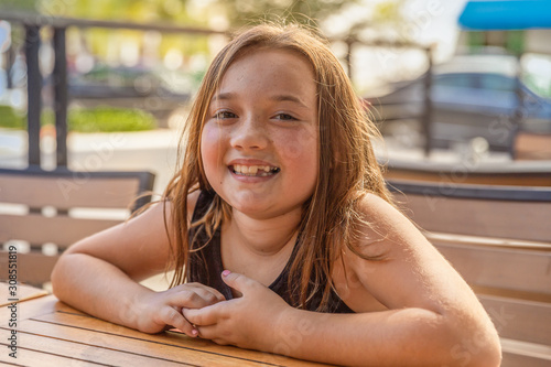 Tablou pe pânză A little girl smiles at the camera while sitting at a table outside on the cafe patio