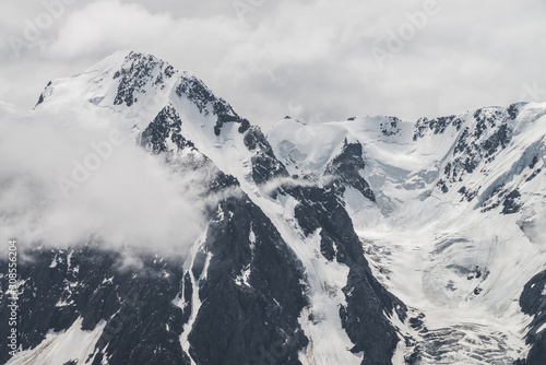 Atmospheric minimalist alpine landscape of big snowy mountain with massive glacier. Low cloud among great rocky mountains. Glacier tongue near snowbound mountainside. Majestic scenery on high altitude