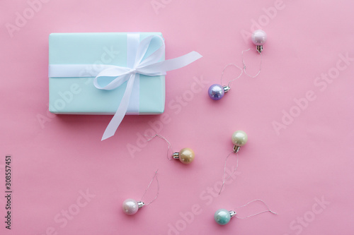 a gift in a blue box tied with a white ribbon on a pink paper background sits next to small shiny Christmas balls of loop shades