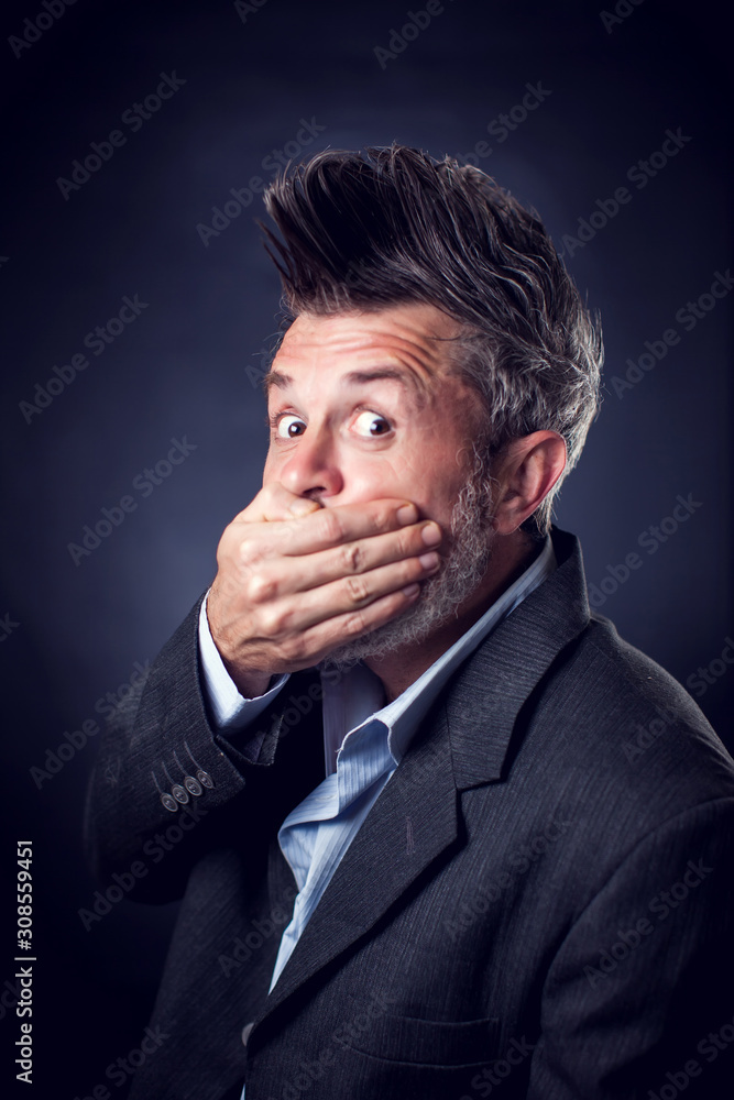 A portrait of shocked bearded man with iroquois wearing suit closed mouth with hands. People and emotions concept