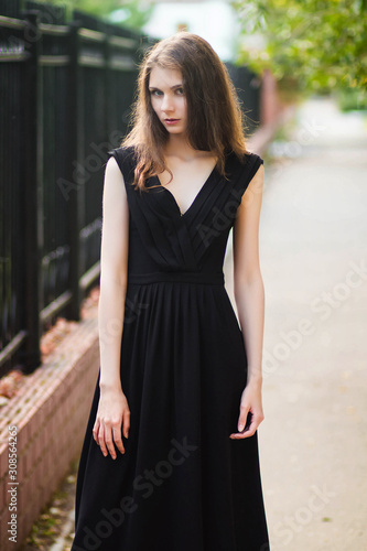 young, beautiful girl in black dress, outdoor photo on a background of steel fence, summer evening. Moddel with clean skin.