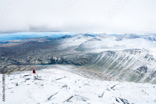 Girl with red hooded jacket and backpack are hiking in snow covered mountains with beautiful scenic landscape view and foggy weather. Scenery, lifestyle, active and outdoor concept.