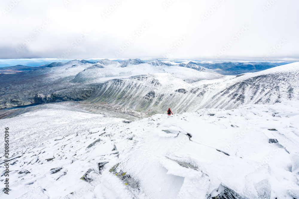 Obraz premium Girl with red hooded jacket and backpack are hiking in snow covered mountains with beautiful scenic landscape view and foggy weather. Scenery, lifestyle, active and outdoor concept.