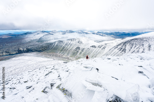 Fototapeta Naklejka Na Ścianę i Meble -  Girl with red hooded jacket and backpack are hiking in snow covered mountains with beautiful scenic landscape view and foggy weather. Scenery, lifestyle, active and outdoor concept.
