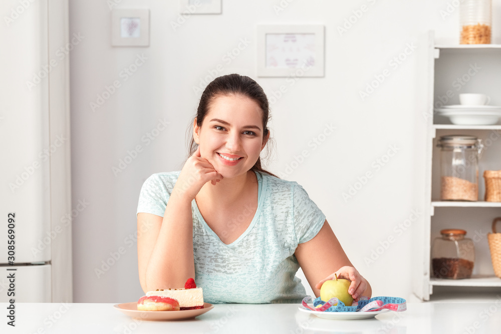 Making Healthy Choice. Chubby girl sitting at kitchen with desserts and plate with apple and tape measure looking camera smiling cheerful Making Healthy Choice. Chubby girl sitting at kitchen with desserts and plate with apple and tape measure looking camera smiling cheerful