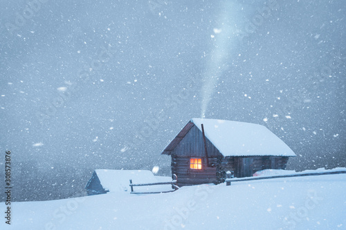 Fantastic winter landscape with wooden house in snowstorm in snowy mountains. Smoke comes from the chimney of snow covered hut. Christmas holiday concept