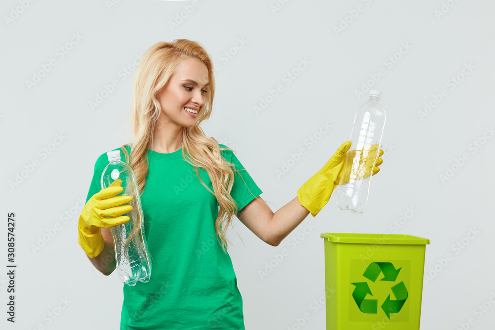 A young woman volunteer in green T-shirts and gloves collects and throws plastic waste into a container for recycling. White studio background A young woman volunteer in green T-shirts and gloves collects and throws plastic waste into a container for recycling. White studio background
