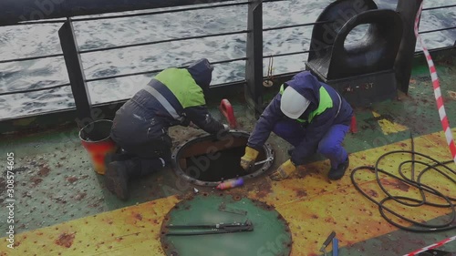 professional mechanics in uniform unscrew pins of cargo tank hatch with spanners on oil tanker deck in open sea photo