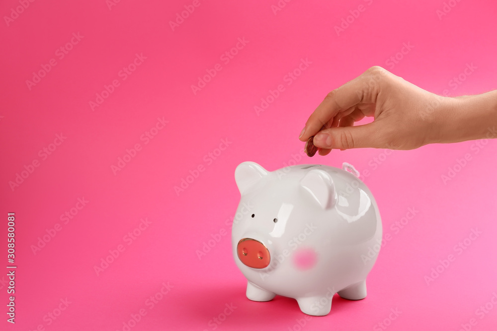 Woman putting coin into piggy bank on pink background, closeup. Space for text