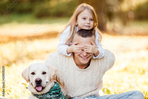 Happy father and daughter with dog labrador is having fun are sitting on green grass in park. © zadorozhna