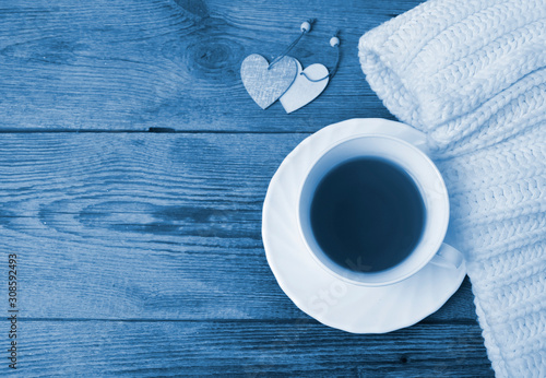 Cozy flatlay composition with white knitted scarf, cup of tea, two decorative heart on wooden desk. Christmas, Valentines day tredy color blue concept. Copyspace.