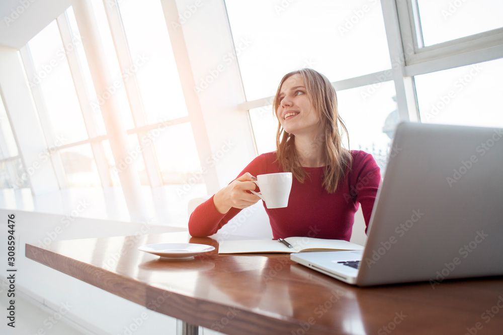 Obraz premium young girl freelancer sitting with a laptop in a cafe working and drinking coffee, student use a computer in the morning near the window