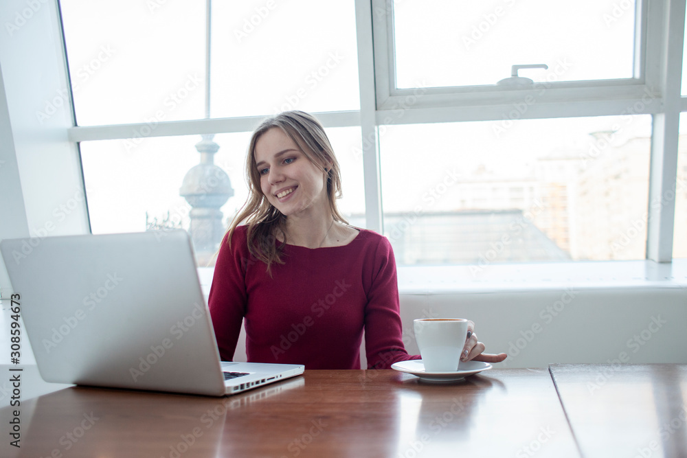 young girl freelancer sitting with a laptop in a cafe working and drinking coffee, student use a computer in the morning near the window young girl freelancer sitting with a laptop in a cafe working and drinking coffee, student use a computer in the morning near the window