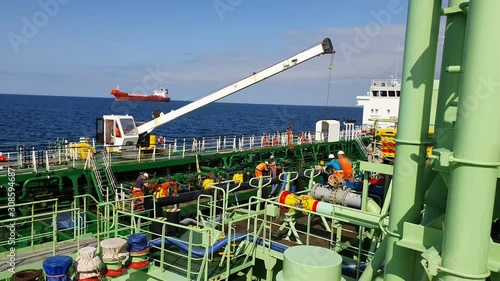 white hoist lowers down to help sailors setting bunker hoses on deck against calm sea with red tanker on raid