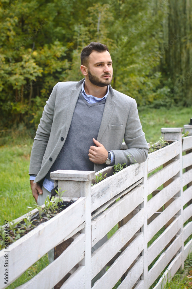 Fototapeta premium Portrait of a sexy man dressed in a suit. Fashionable guy in the autumn nature near a white wooden fence. Businessman relaxes. Skok photo for design