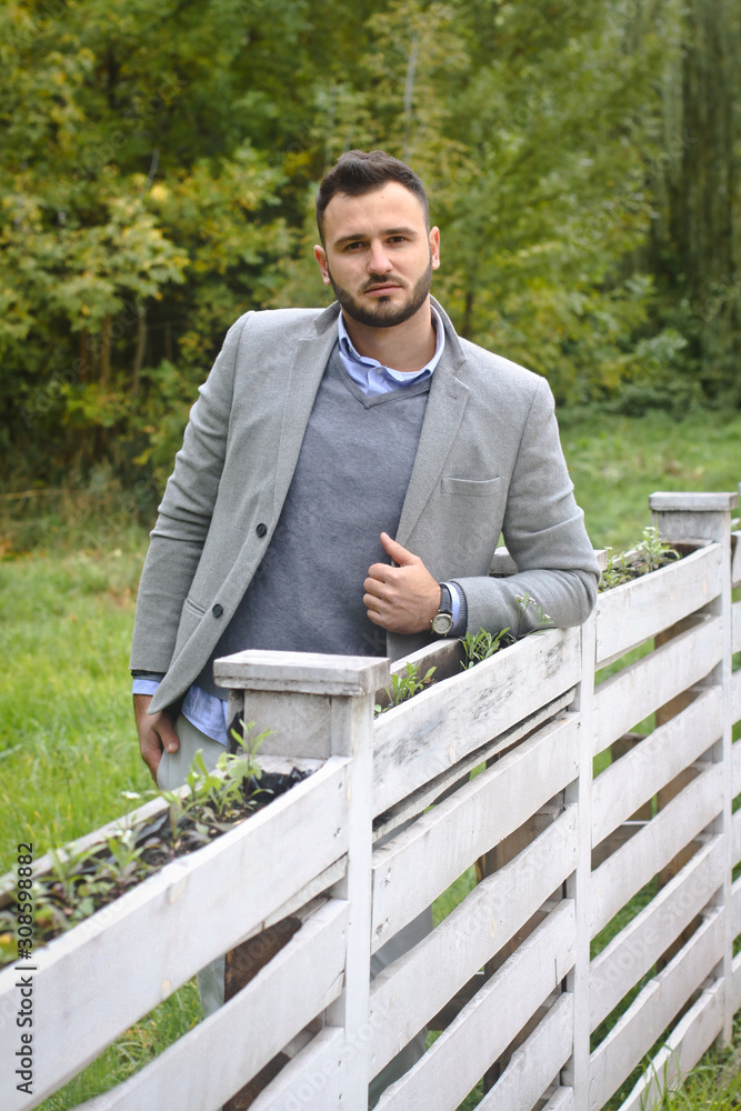 Fototapeta premium Portrait of a sexy man dressed in a suit. Fashionable guy in the autumn nature near a white wooden fence. Businessman relaxes. Skok photo for design