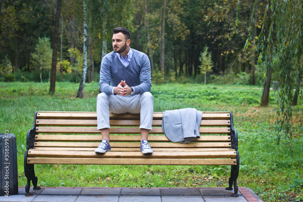 Young sexy guy sitting on a bench in an autumnal, summer park. A man contemplating new ideas while walking. European countryside. Stock Photo Young sexy guy sitting on a bench in an autumnal, summer park. A man contemplating new ideas while walking. European countryside. Stock Photo