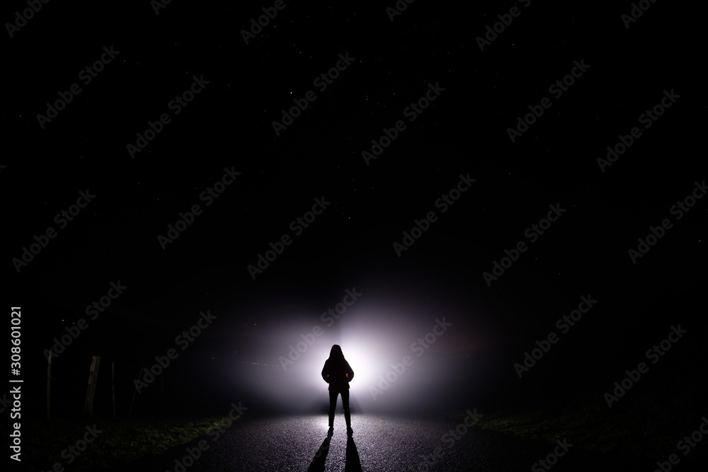 Fototapeta premium Sylwetka kobiety w ciemności. Fotografia nocna. Jasne światło świecące za ciemną tajemniczą postacią. Widmowa, mistyczna, surrealistyczna osoba stojąca.