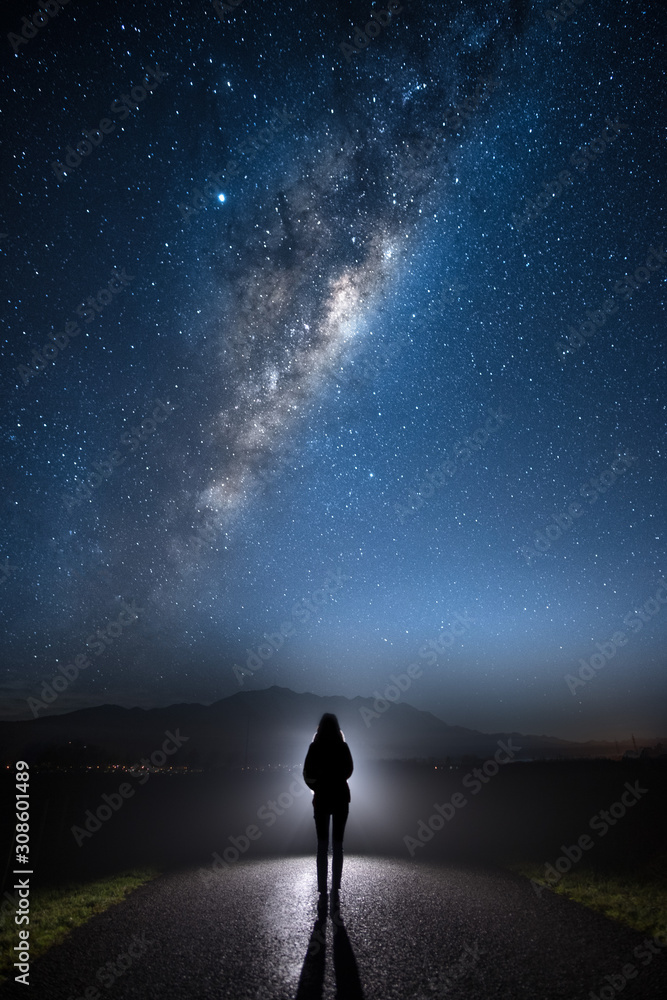 A mysterious lost person standing in the middle of the road looking into bright light with milky way starry night sky. A mysterious lost person standing in the middle of the road looking into bright light with milky way starry night sky.