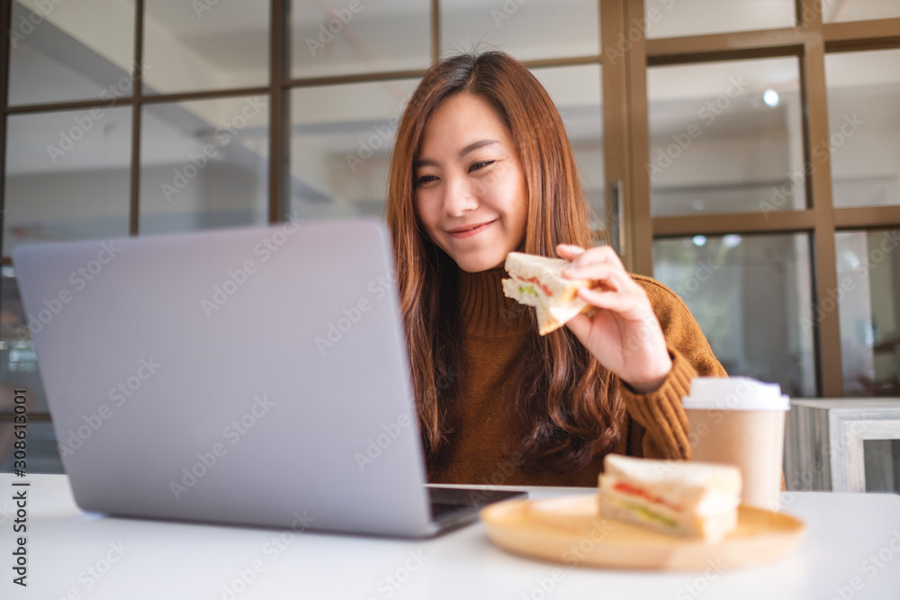 An asian woman holding and eating whole wheat sandwich while working on laptop computer An asian woman holding and eating whole wheat sandwich while working on laptop computer