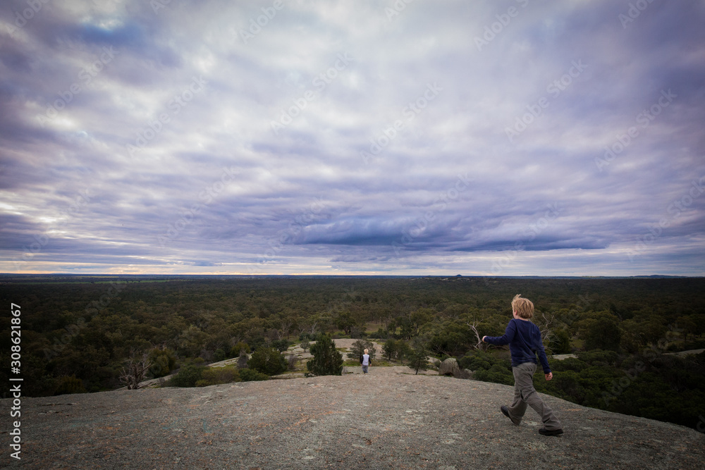 Boy walking along large rock with sweeping view of the Terrick Terrick National Park on an overcast day Boy walking along large rock with sweeping view of the Terrick Terrick National Park on an overcast day