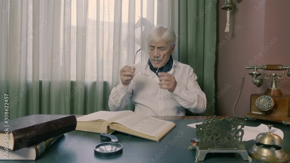 A gray-haired man at age, a writer in a white shirt, thinks about his work and looks for the necessary information in a book lying on a comfortable desk in retro style in his office A gray-haired man at age, a writer in a white shirt, thinks about his work and looks for the necessary information in a book lying on a comfortable desk in retro style in his office