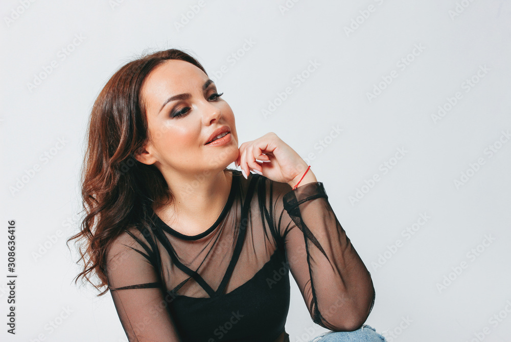 Smiling charming brunette young woman with bright makeup dressed in black transparent blouse and fashionable ripped jeans sitting on white background isolated Smiling charming brunette young woman with bright makeup dressed in black transparent blouse and fashionable ripped jeans sitting on white background isolated