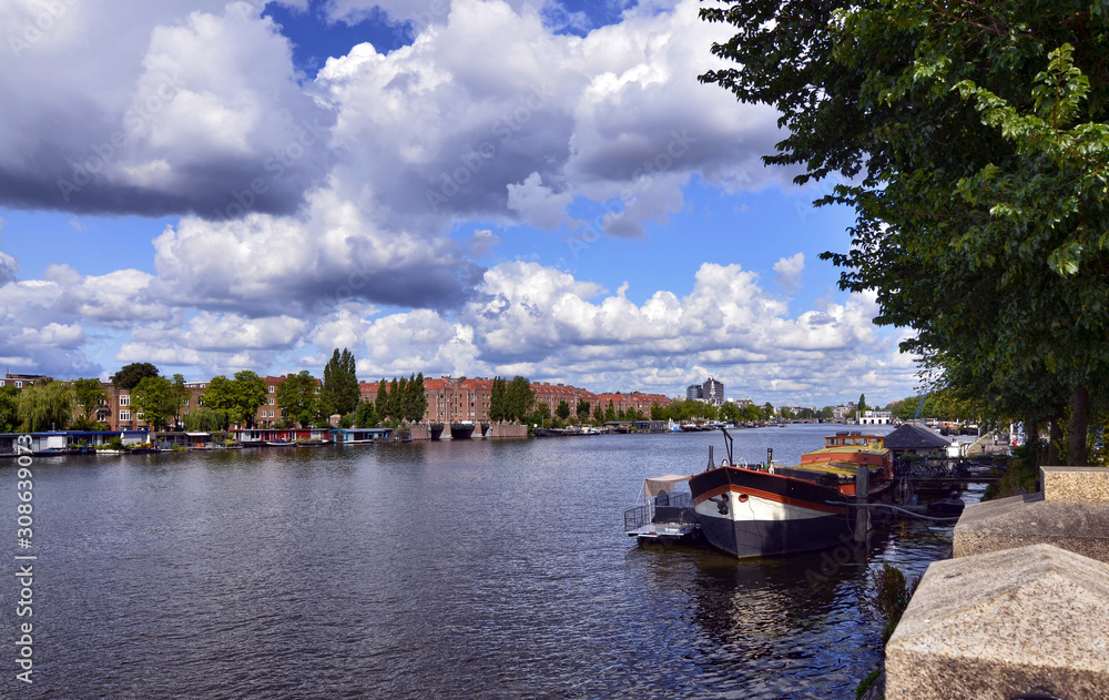 Fototapeta premium Amsterdam, Holland, August 2019. View of the Amstel River, outskirts of the city. A large moored boat is used as a houseboat. Sunny day with blue sky and white clouds.