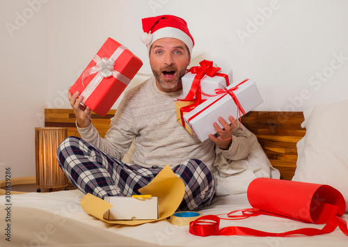 packing Chirstmas presents   young happt and attractive man sitting on bed in Santa Claus hat wrapping and preparing xmas gifts and boxes with paper and tape smiling excited