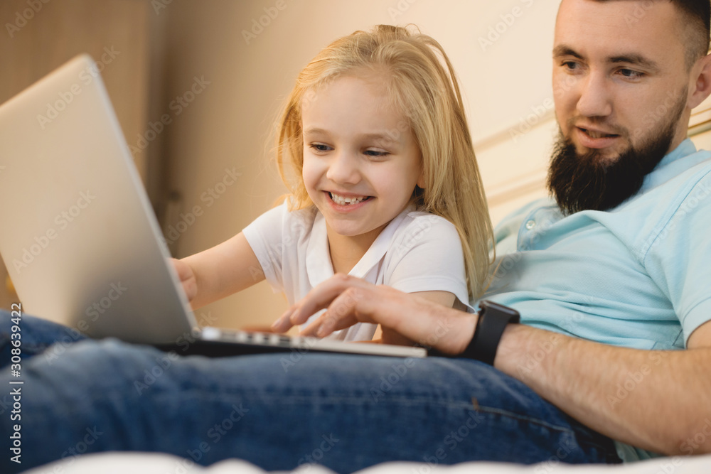 Young bearded father and his little daughter looking to a laptop screen while leaning on a bed at home. Young bearded father and his little daughter looking to a laptop screen while leaning on a bed at home.