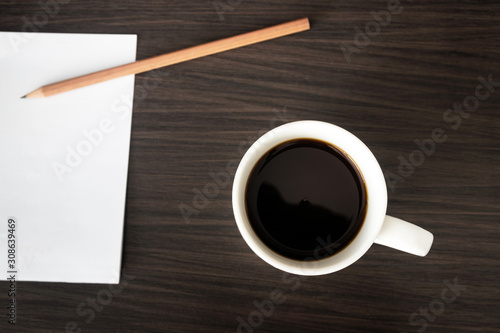 Black coffee cup with blank notepad and pencil on dark brown wooden table. Overhead shot