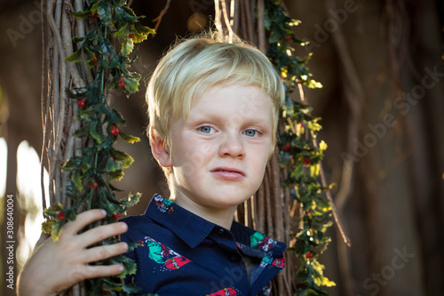 Lovely close up portrait of young boy wearning Christmas shirt resting head against fig tree roots covered in mistletoe photo