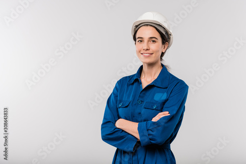 Happy young cross-armed female engineer in blue workwear and hardhat
