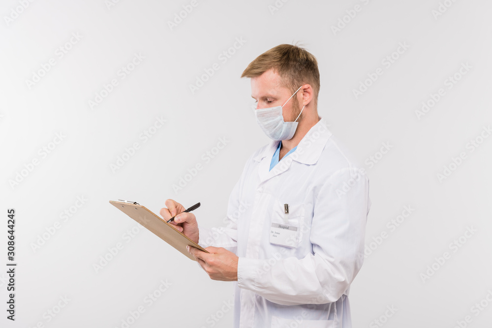 Young doctor in whitecoat and protective mask making prescriptions for patient Young doctor in whitecoat and protective mask making prescriptions for patient