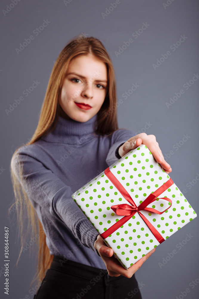 Young caucasian lady with long hair is giving a gift box to you and me isolated on a gray background. Young caucasian lady with long hair is giving a gift box to you and me isolated on a gray background.
