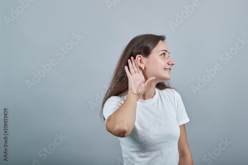 Young lady in white t-shirt over isolated wall holds her palm near her ear and listens to gossips