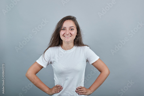 Teen girl standing over isolated gray wall and smiling, she has her hands on her waist. Young woman dressed in white t-shirt