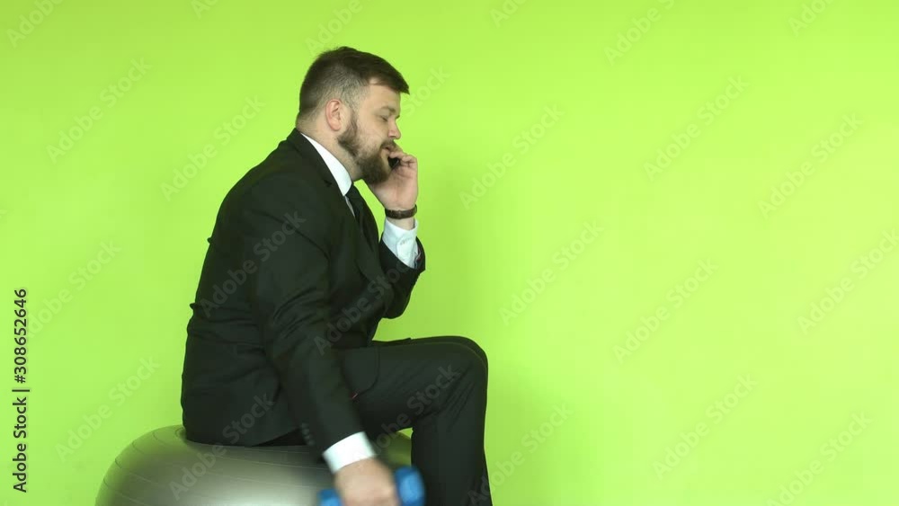 A caucasian male businessman with a beard in a black suit sits on a fitness ball and goes in with a dumbbell. Talking on the phone and sports, copy space A caucasian male businessman with a beard in a black suit sits on a fitness ball and goes in with a dumbbell. Talking on the phone and sports, copy space