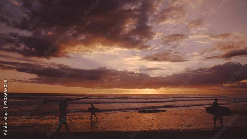Beautiful sunset on a wide and remote sandy Pacific beach at Santa Teresa, Costa Rica. Clouds are colorizing the entire sky and the waves. Two surfers are walking along the shore with their boards. Beautiful sunset on a wide and remote sandy Pacific beach at Santa Teresa, Costa Rica. Clouds are colorizing the entire sky and the waves. Two surfers are walking along the shore with their boards.