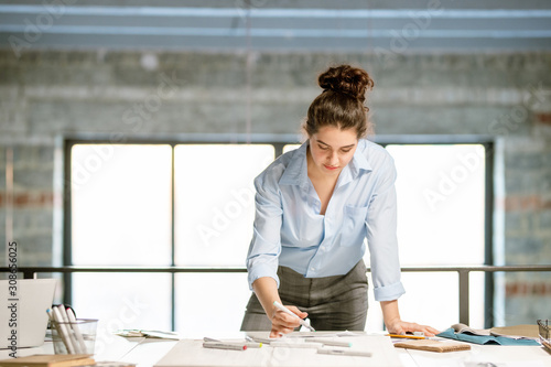 Young serious female designer bending over fashion sketches on desk