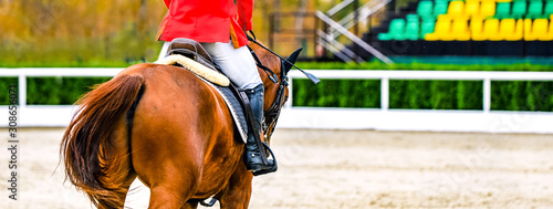 Sorrel dressage horse and rider in red uniform performing jump at show jumping competition. Equestrian sport background. Chesnut horse portrait during dressage competition.