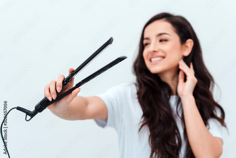 Pretty, smiling girl holding a straightener in her hands. Closeup, blurred background Pretty, smiling girl holding a straightener in her hands. Closeup, blurred background