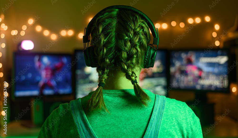 Obraz premium Gamer or streamer girl at home in a dark room playing video games with friends online. Young man sitting in front of three monitors, green creative light